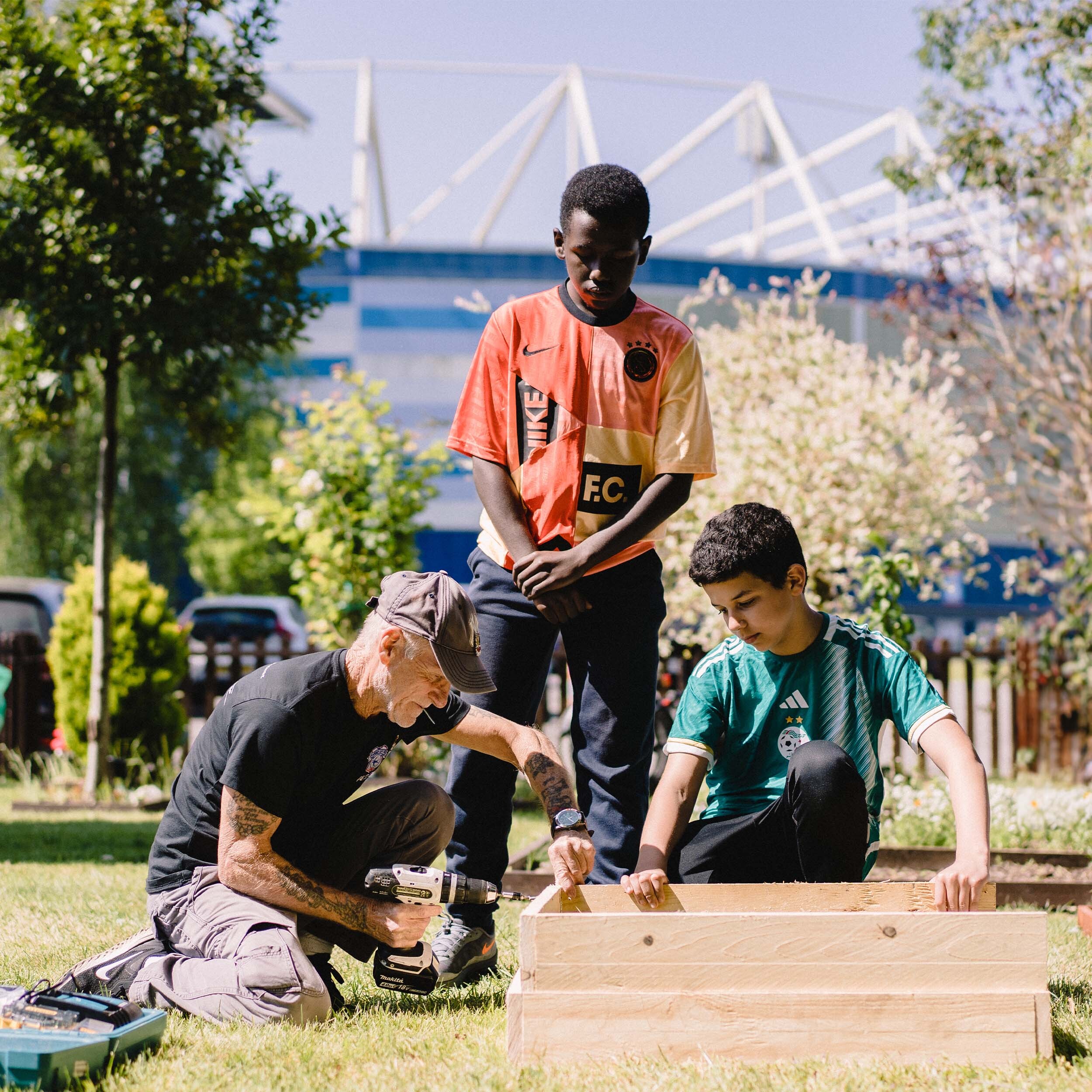 an older man teaching two young males how to build something out of wood, outside cardiff city stadium