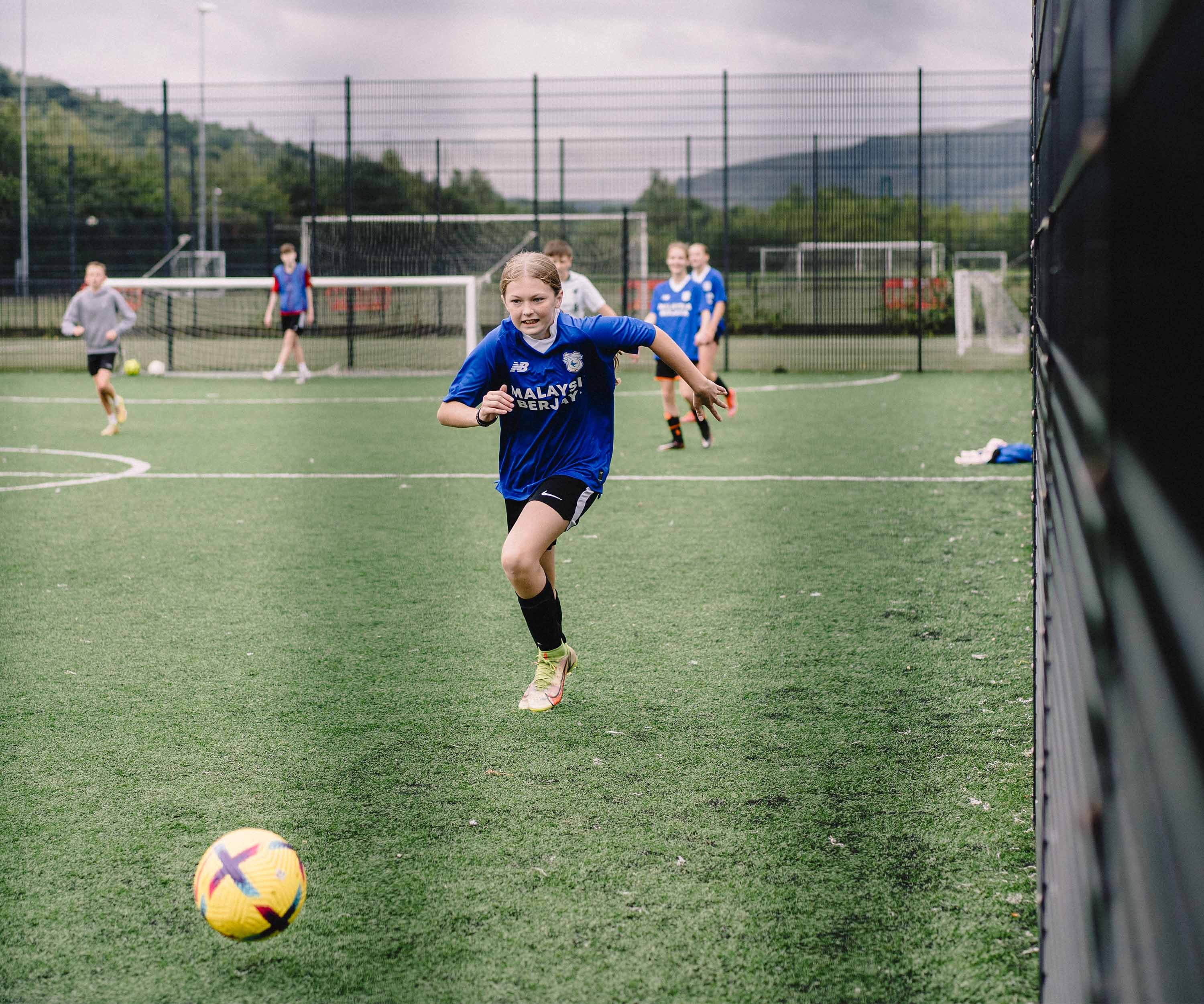 a young girl playing football in a astroturf court in the community, wearing a cardiff city fc kit