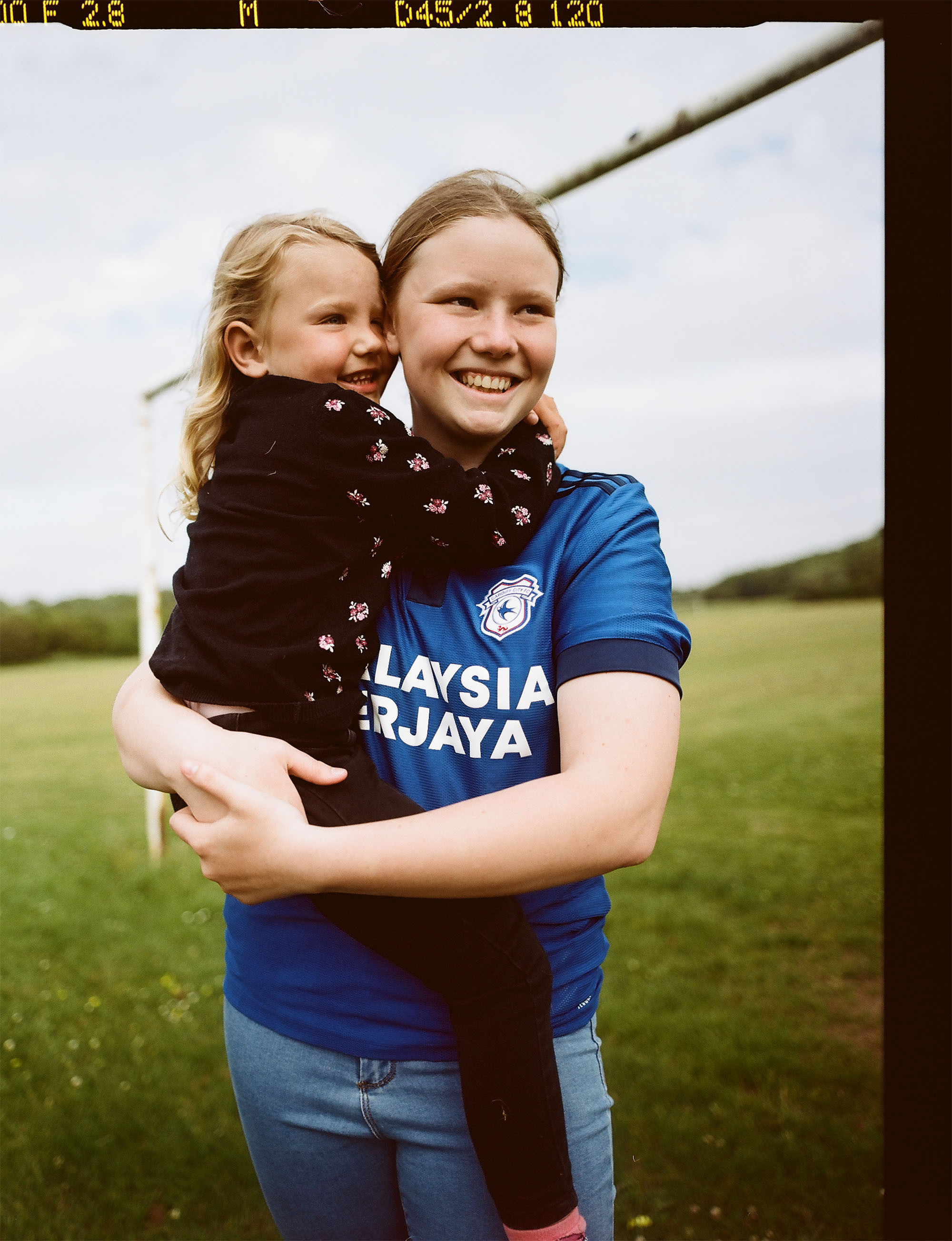 A young girl wearing the 2021/2022 Cardiff City Football Club home shirt. She's holding her younger sister in her arms whilst they both laugh together.