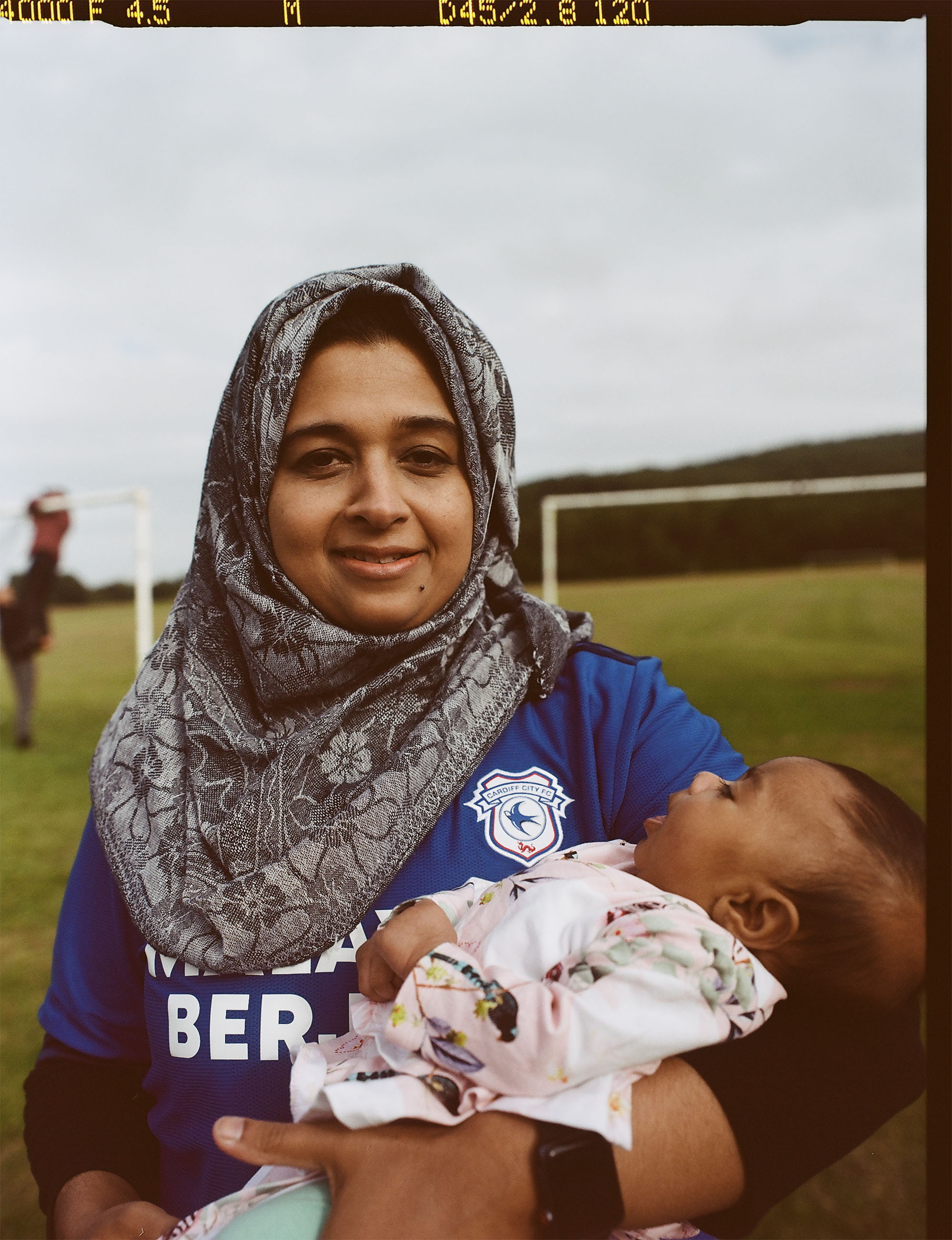 A woman in a head scarf wearing the 2021/2022 Cardiff City Football Club home shirt. She's holding an infant child in her arms.
